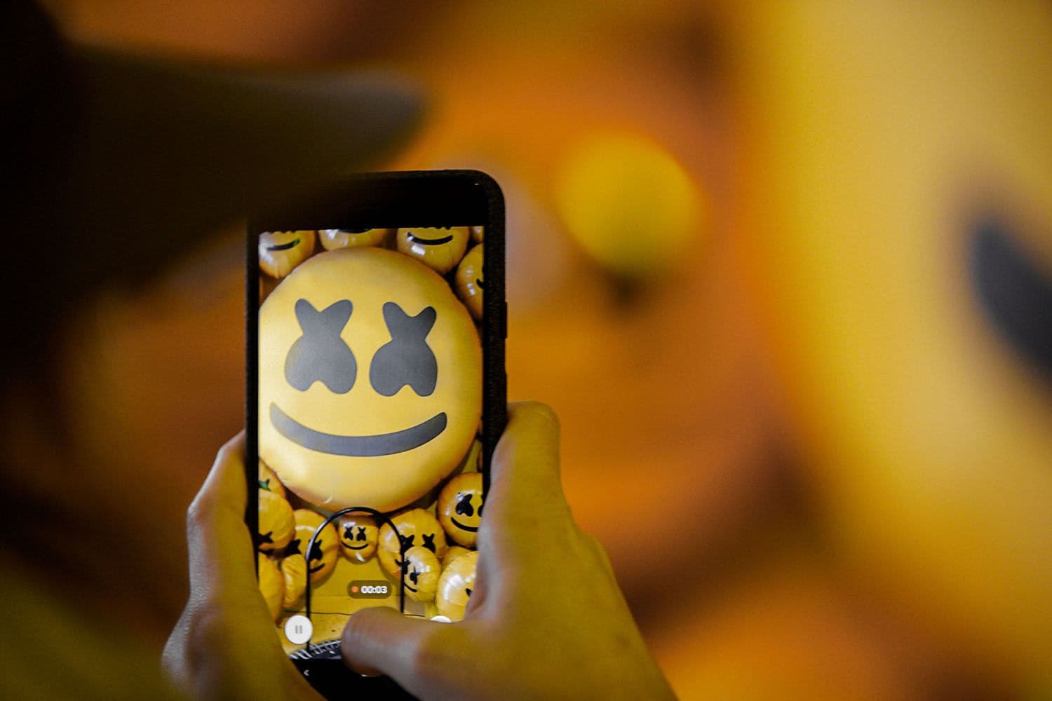 Close-up of a hand holding a phone to photograph a marshmello mask emoji sculpture.