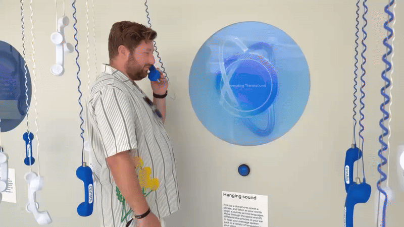 A man holds a blue corded telephone receiver to his ear beside a glowing circular blue screen, surrounded by hanging phone receivers on a wall.