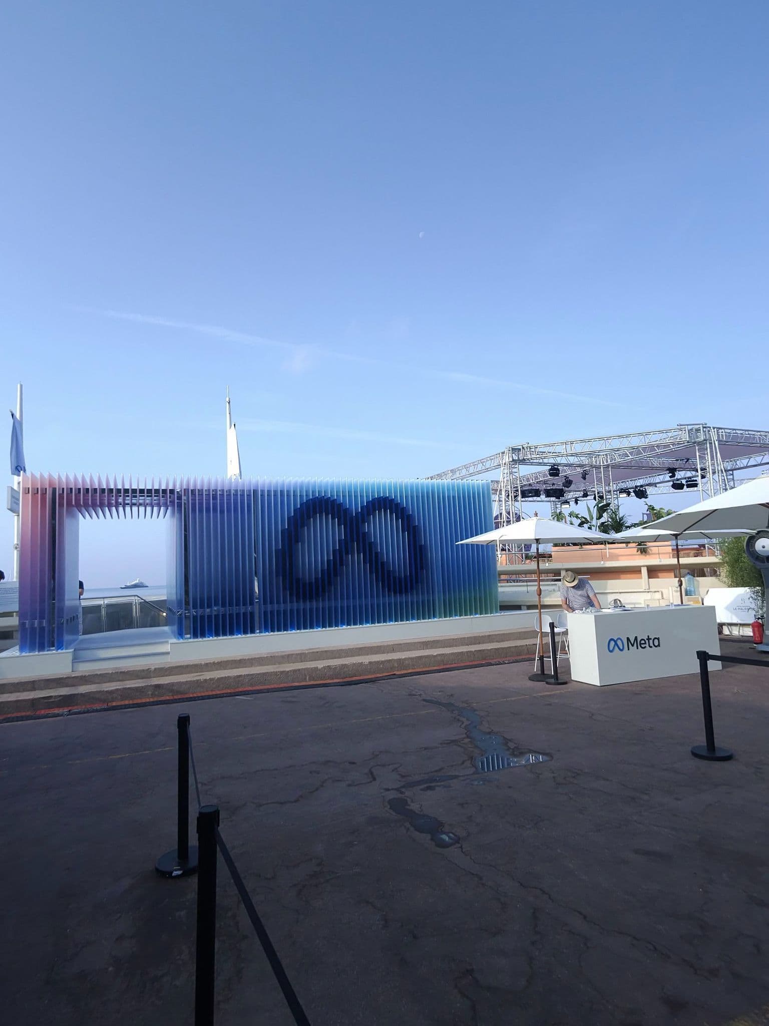 A translucent blue and purple gradient wall with a Meta infinity logo and an open archway, set against a clear sky with a stage structure nearby.
