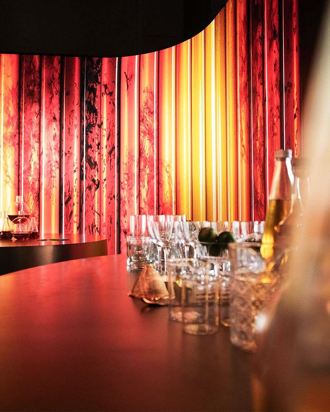 Red-lit lounge area with glassware and bottles on a wooden counter in front of a striped red curtain.