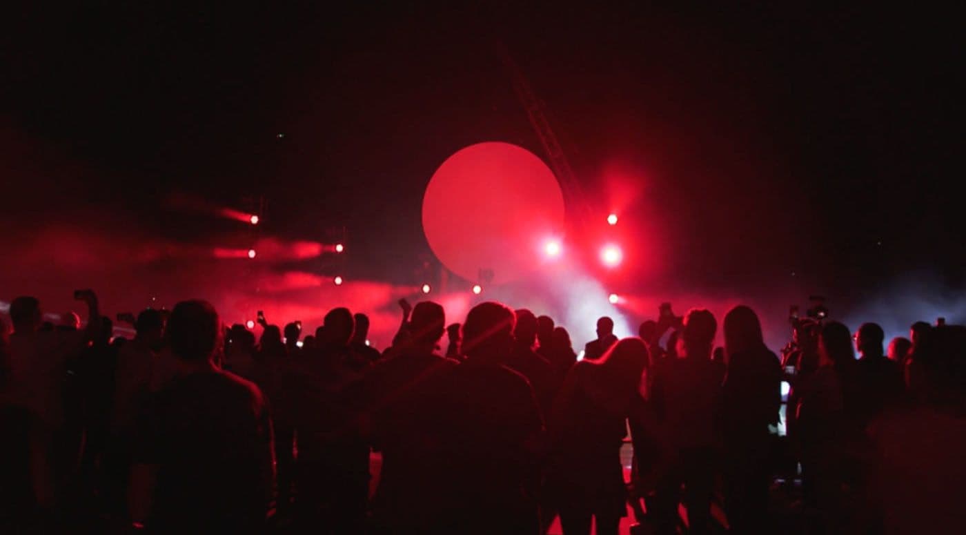 Crowd silhouetted against a large red circular light and stage haze at a night event.
