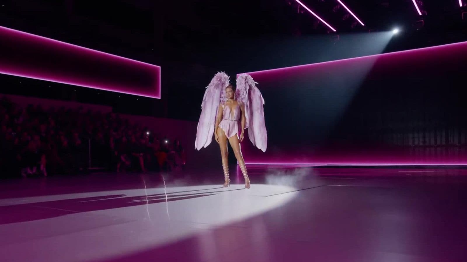 A model wearing pink feathered wings walks the runway framed by vertical pink neon light bars.