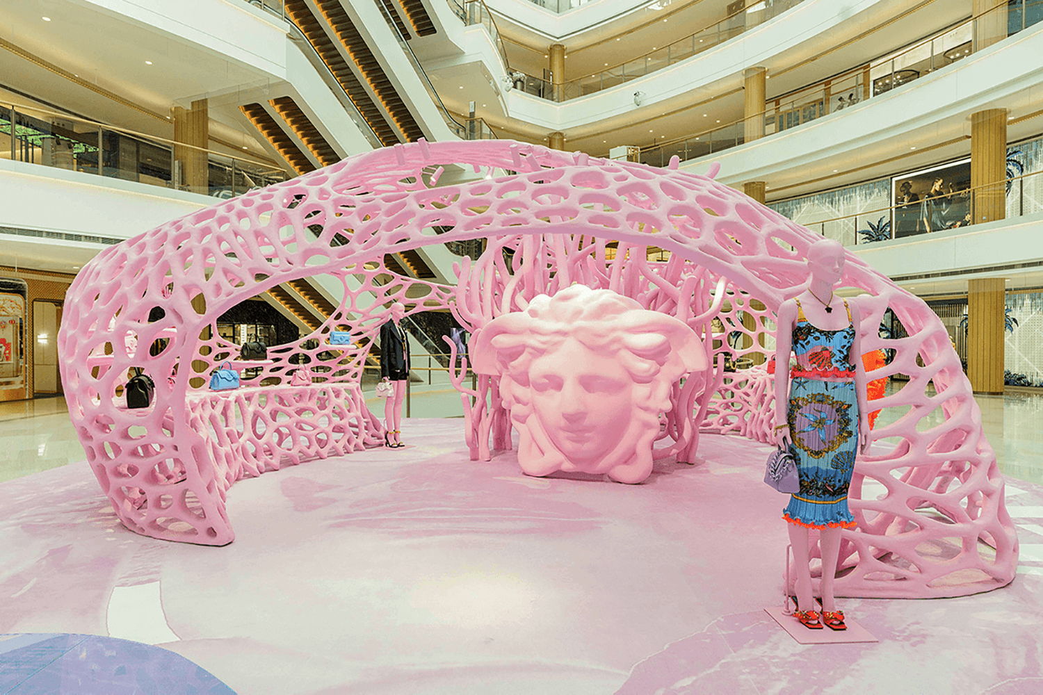 Wide view of the pink lattice archway pavilion in the mall atrium with the Medusa head sculpture at its base and a person in a colorful printed outfit standing beside it.