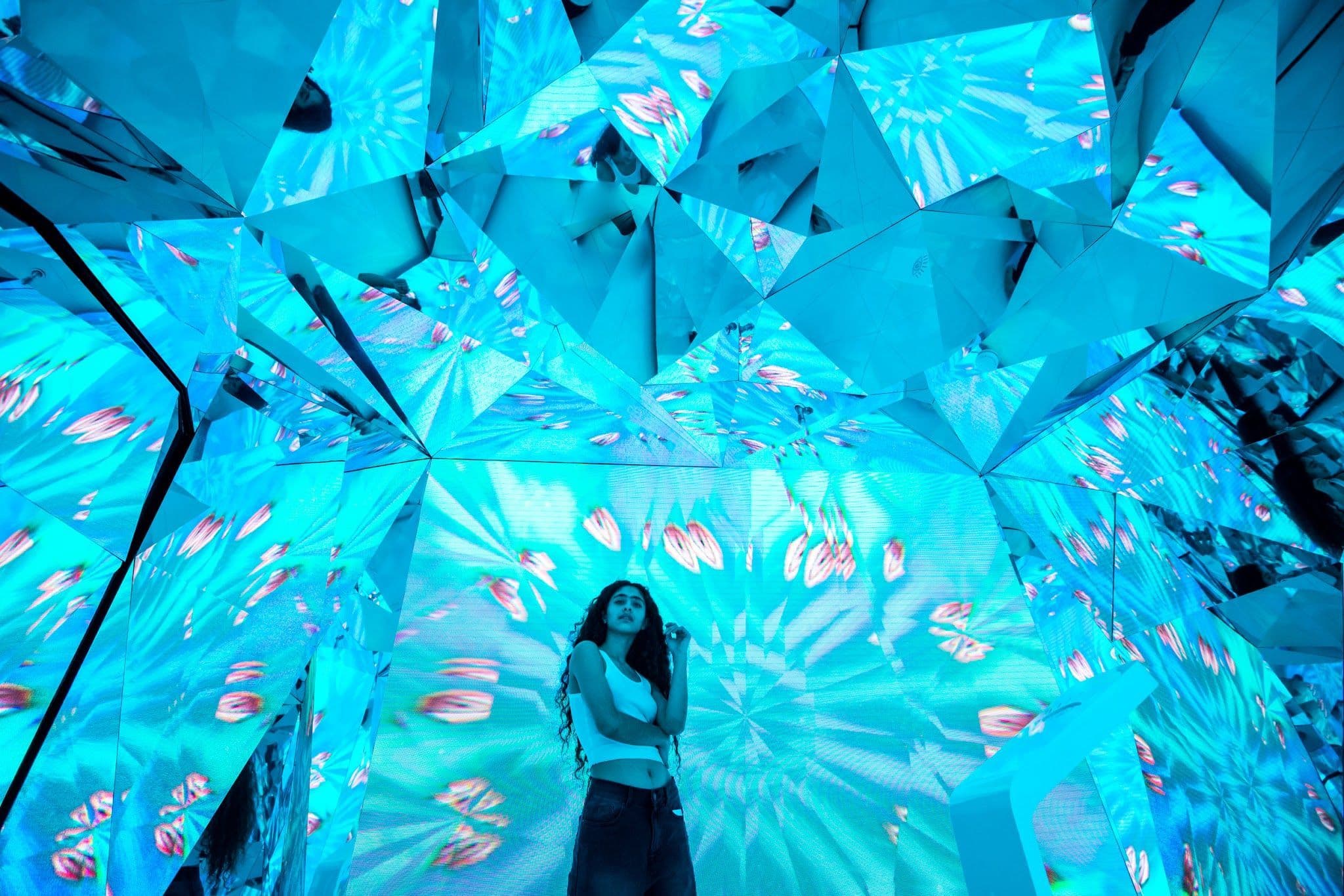 A person stands inside a symmetrical teal and blue kaleidoscopic mirrored room.