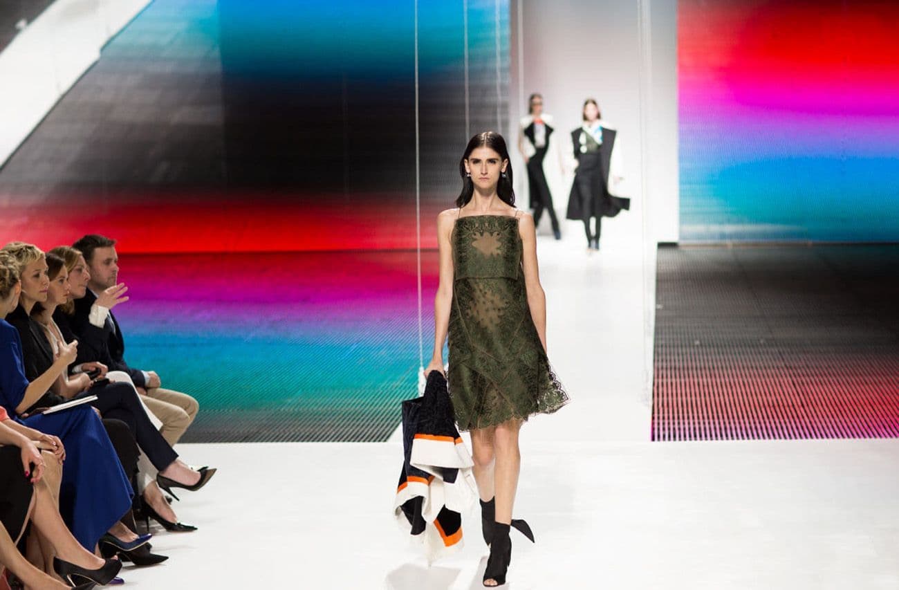 A model in an olive sleeveless dress walks the runway against a dark backdrop with colorful vertical stripes.