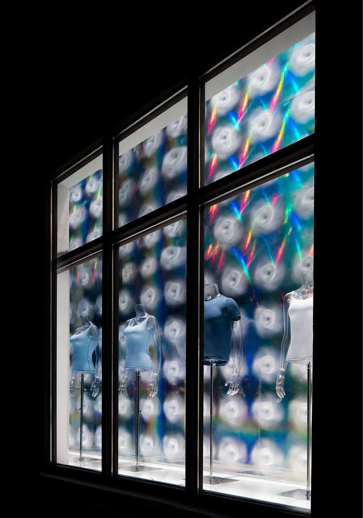Angled view through a storefront window of white pinwheel ornaments and rainbow light streaks above mannequins.