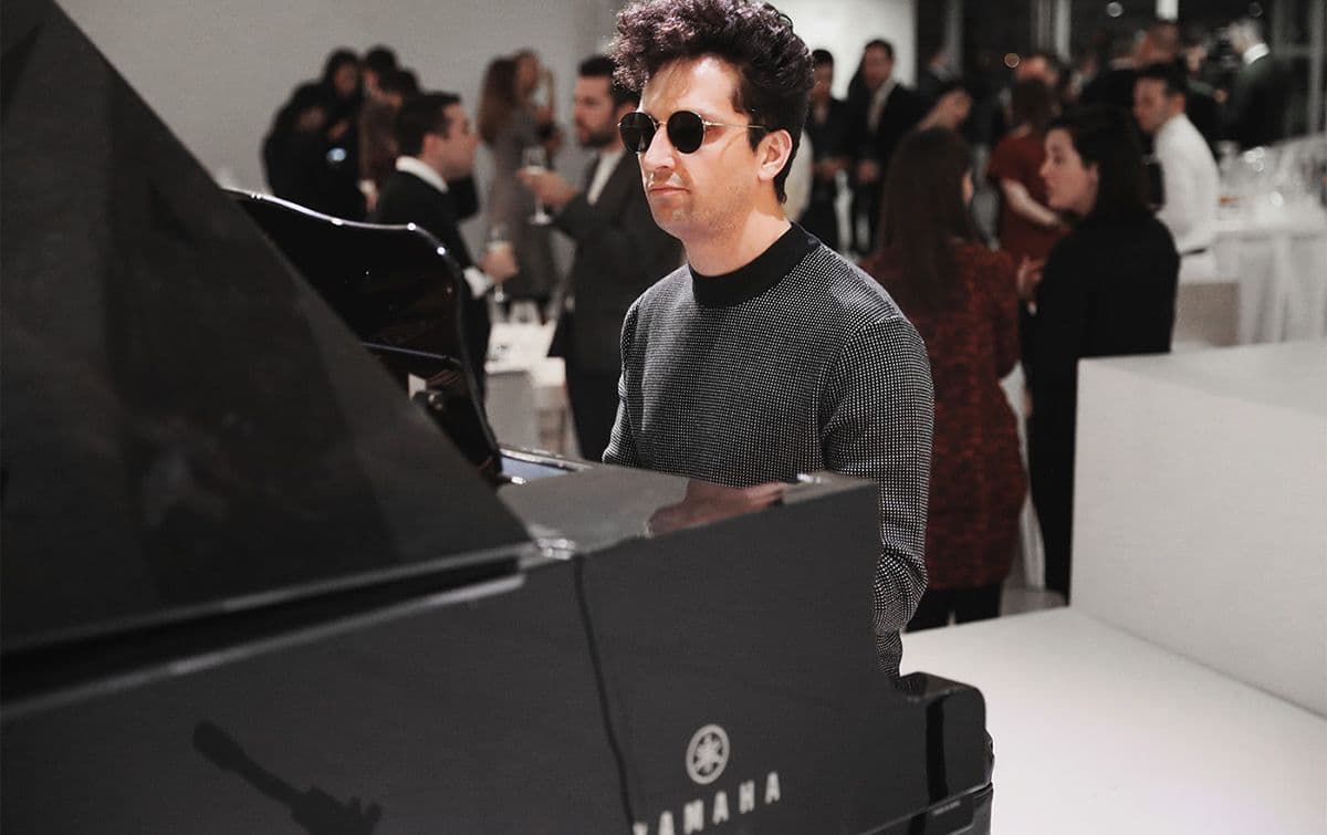 Man wearing sunglasses playing a grand piano at an event with a blurred crowd behind him