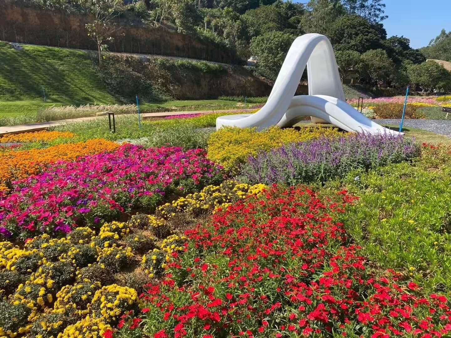 White curved slide sculpture surrounded by beds of red, yellow, purple, and orange flowers.