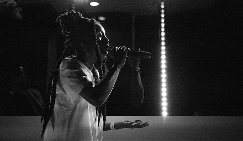 Black-and-white photo of a singer with dreadlocks performing into a microphone, hand raised, on stage.