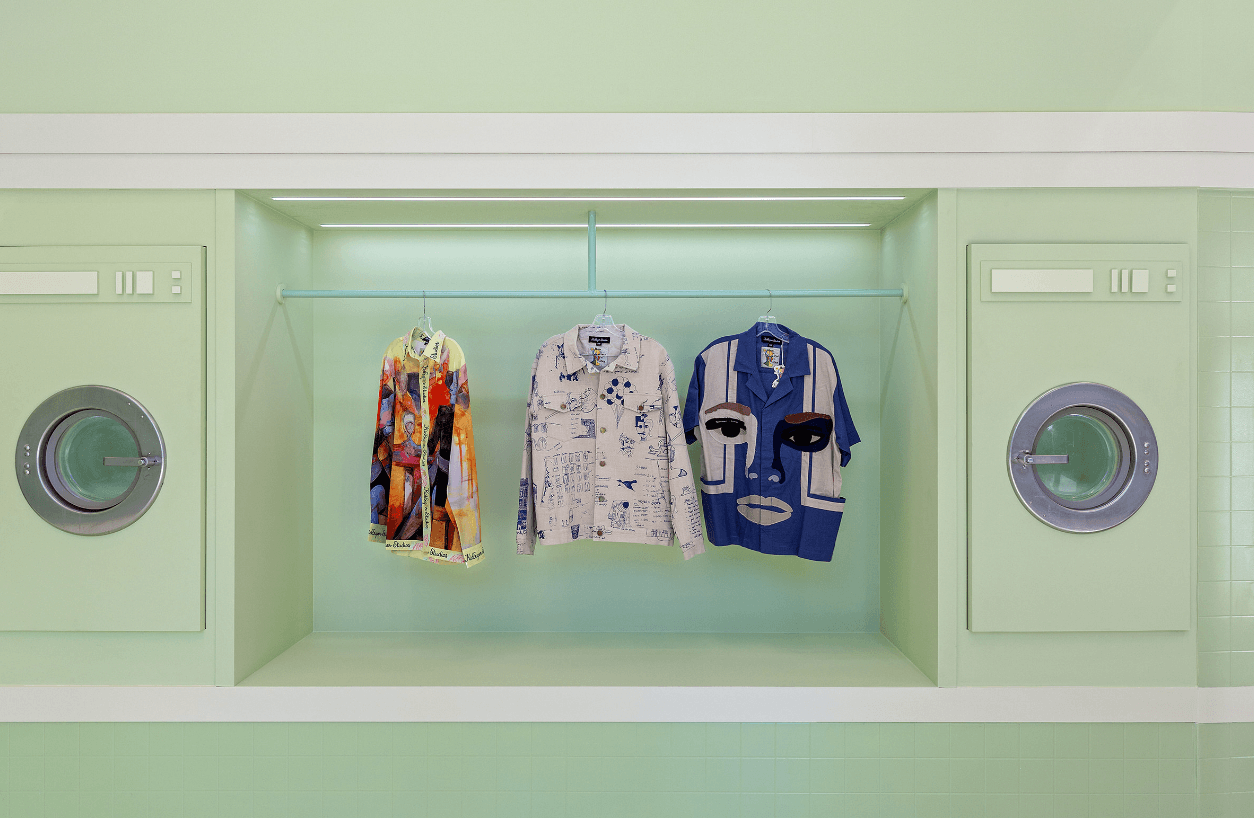 Colorful printed t-shirts and hoodies displayed on shelving between two washing machine units in a mint-green store interior