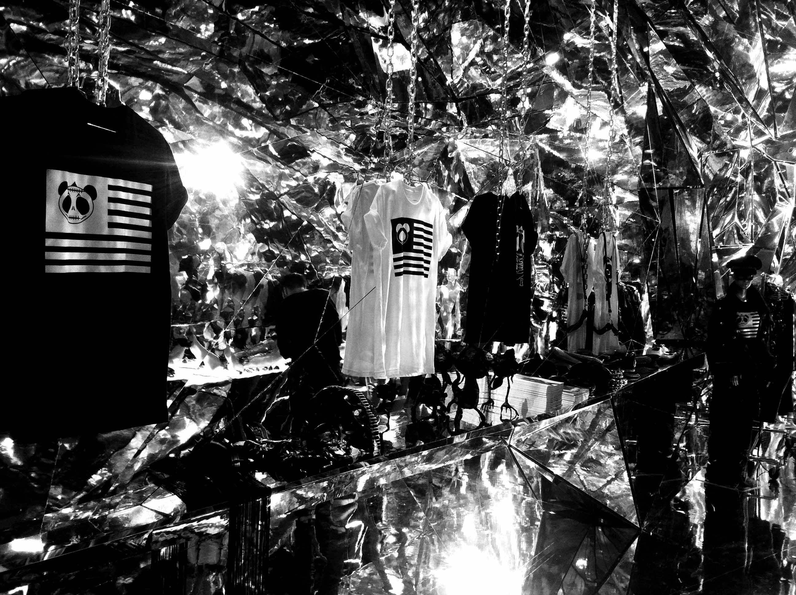 Black and white photo of a shattered glass or foil installation with a clothing rack visible in the background.