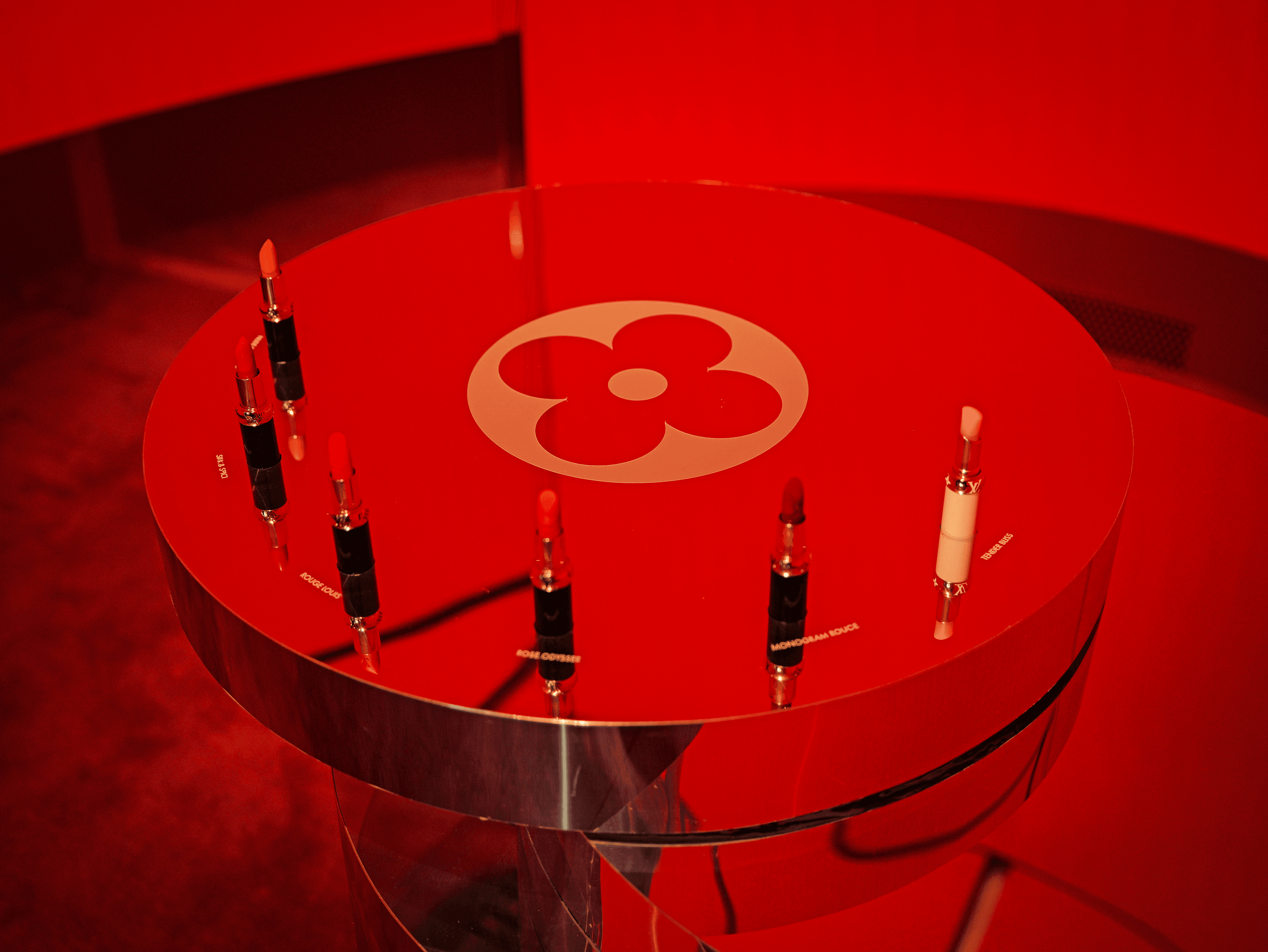 Close-up of a red mirrored round table etched with the Louis Vuitton flower monogram, ringed with lipsticks under red light.