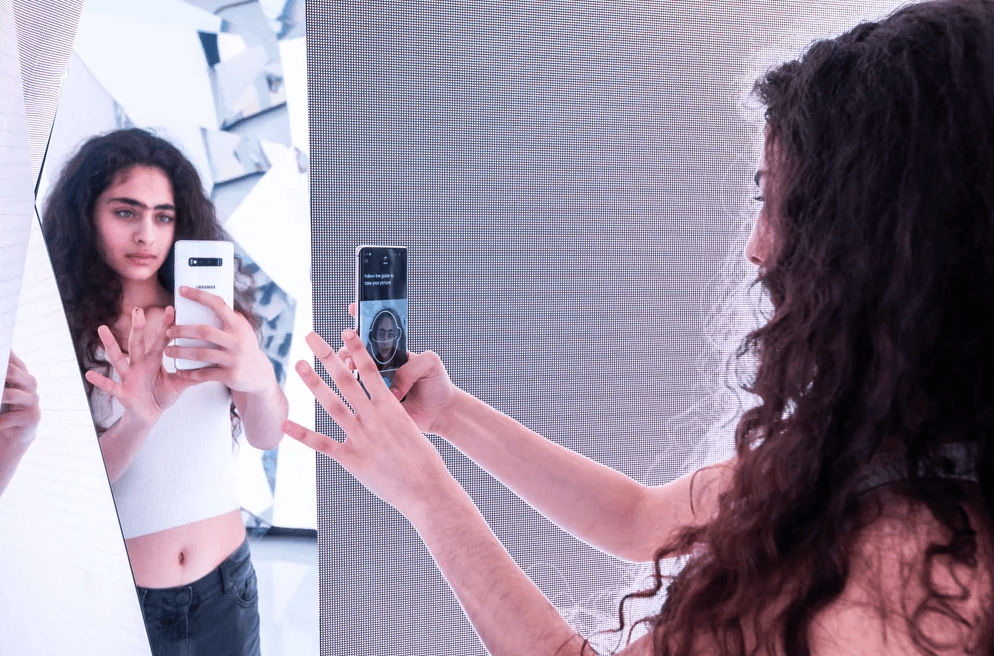 Two women take a selfie with a phone inside a mirrored room with teal-toned reflections.