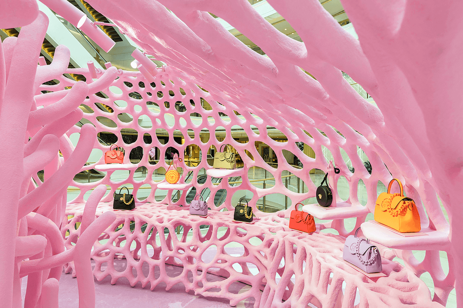 Interior of the pink lattice tunnel with a row of Versace handbags in various colors displayed on shelves along the perforated wall.