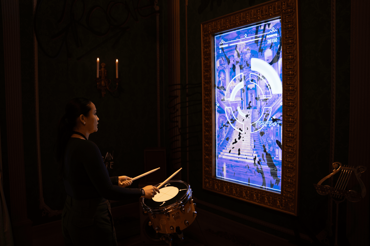 A person plays a snare drum in a dim room beside a glowing blue interactive display framed in gold.