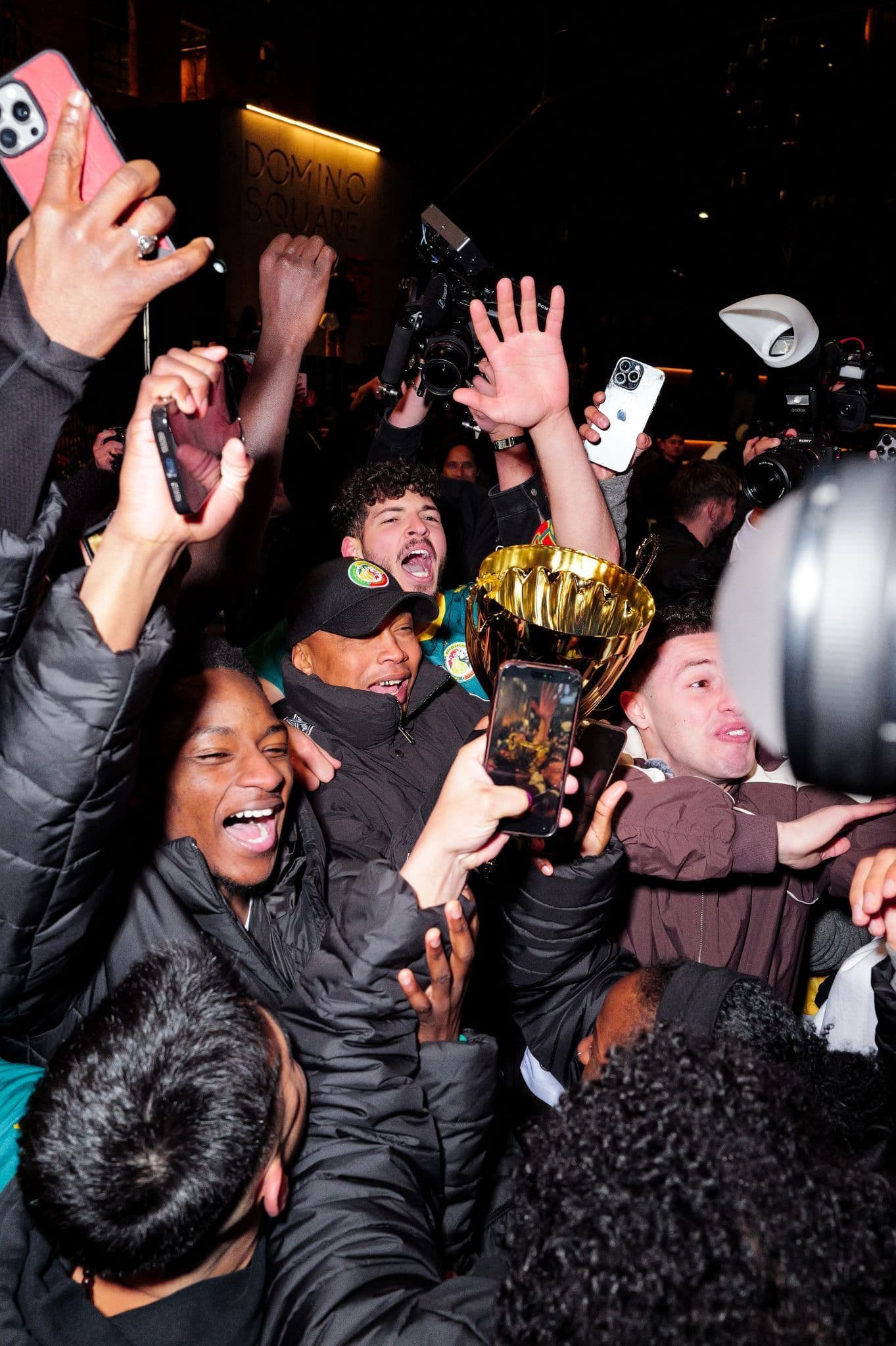 Excited crowd at night raising phones to take photos, some holding up a trophy.