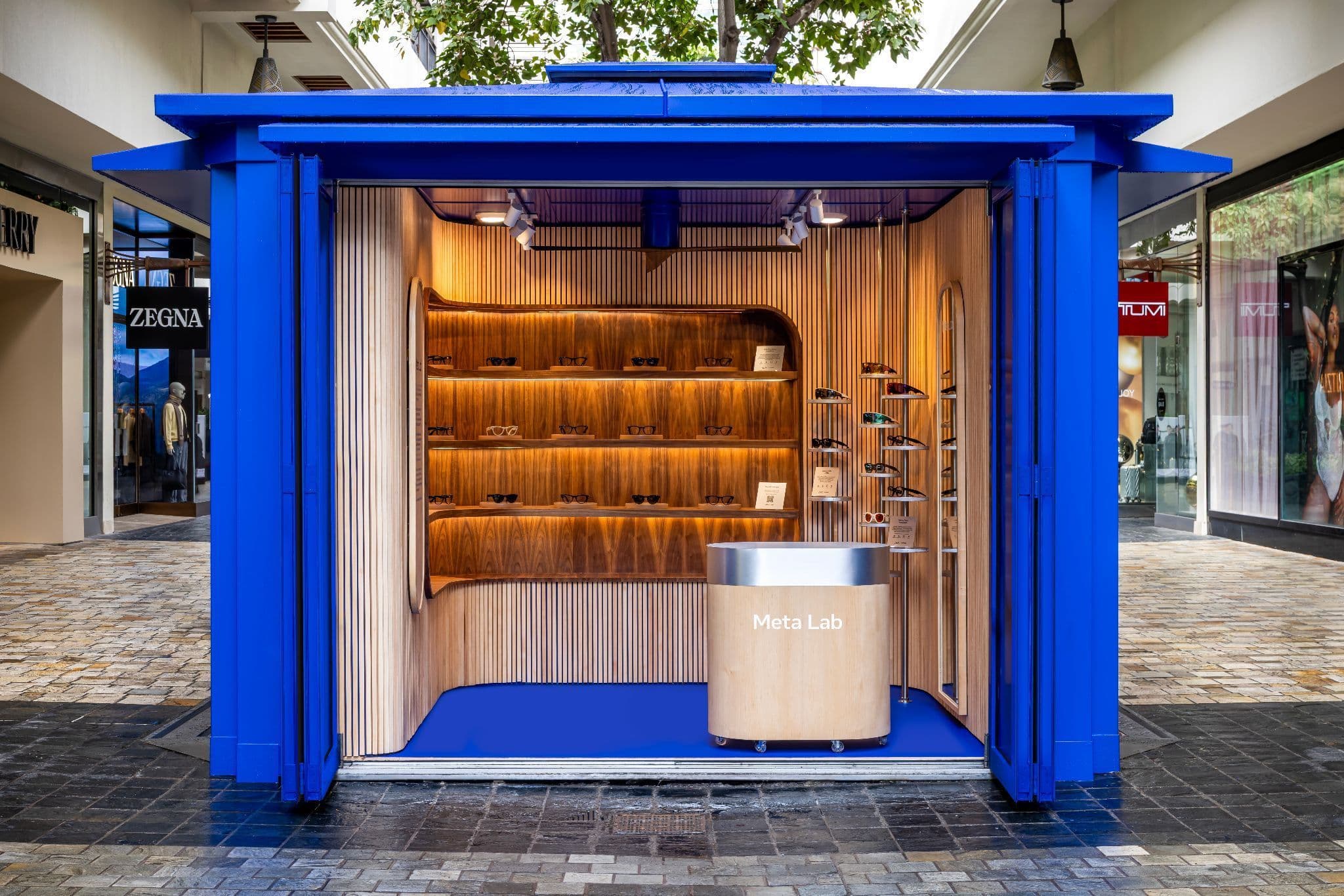 Open blue kiosk with wood shelving and a counter visible through its opened front panel, set in an outdoor shopping plaza.