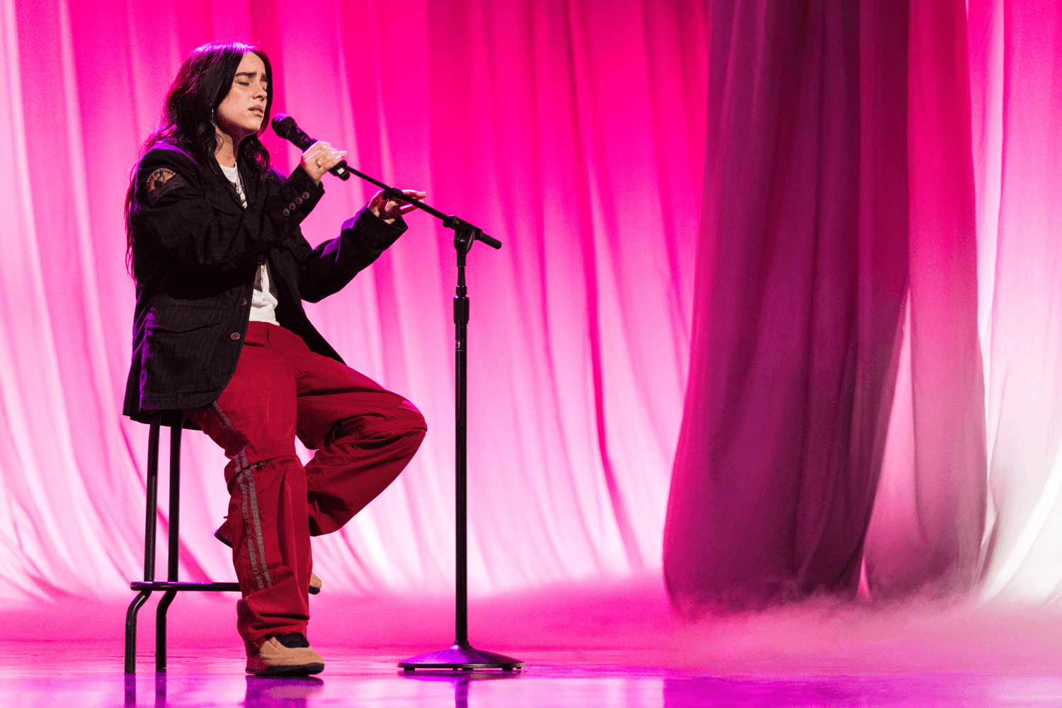 Performer seated on a stool singing into a microphone against a pink curtain backdrop