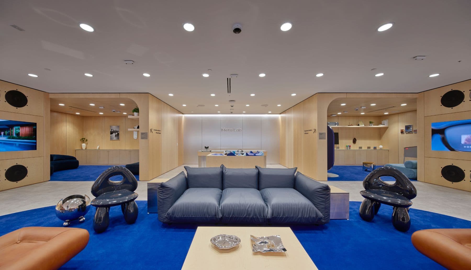Wood-paneled seating area with a gray sofa, patterned ottomans, and wall-mounted screens on either side of a blue carpeted floor.