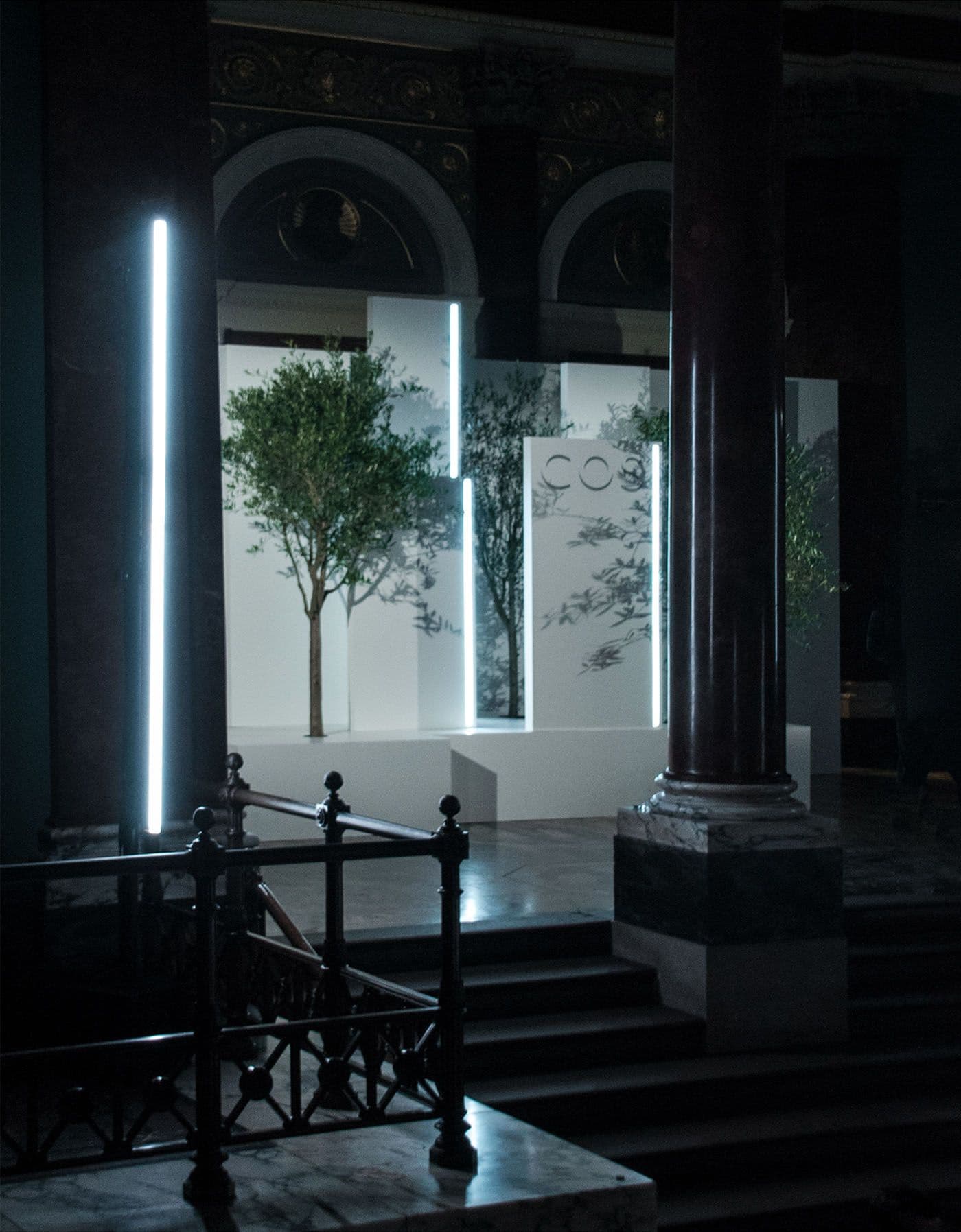 View through a wrought-iron stair railing and stone columns toward a lit olive-tree installation with COS branding.