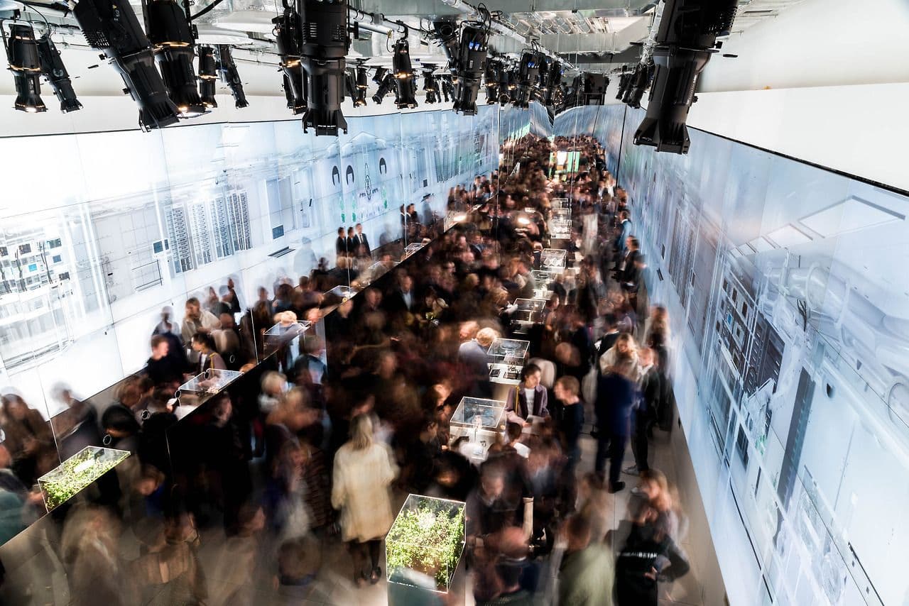 Motion-blurred wide shot of a crowd walking through an illuminated exhibition corridor.