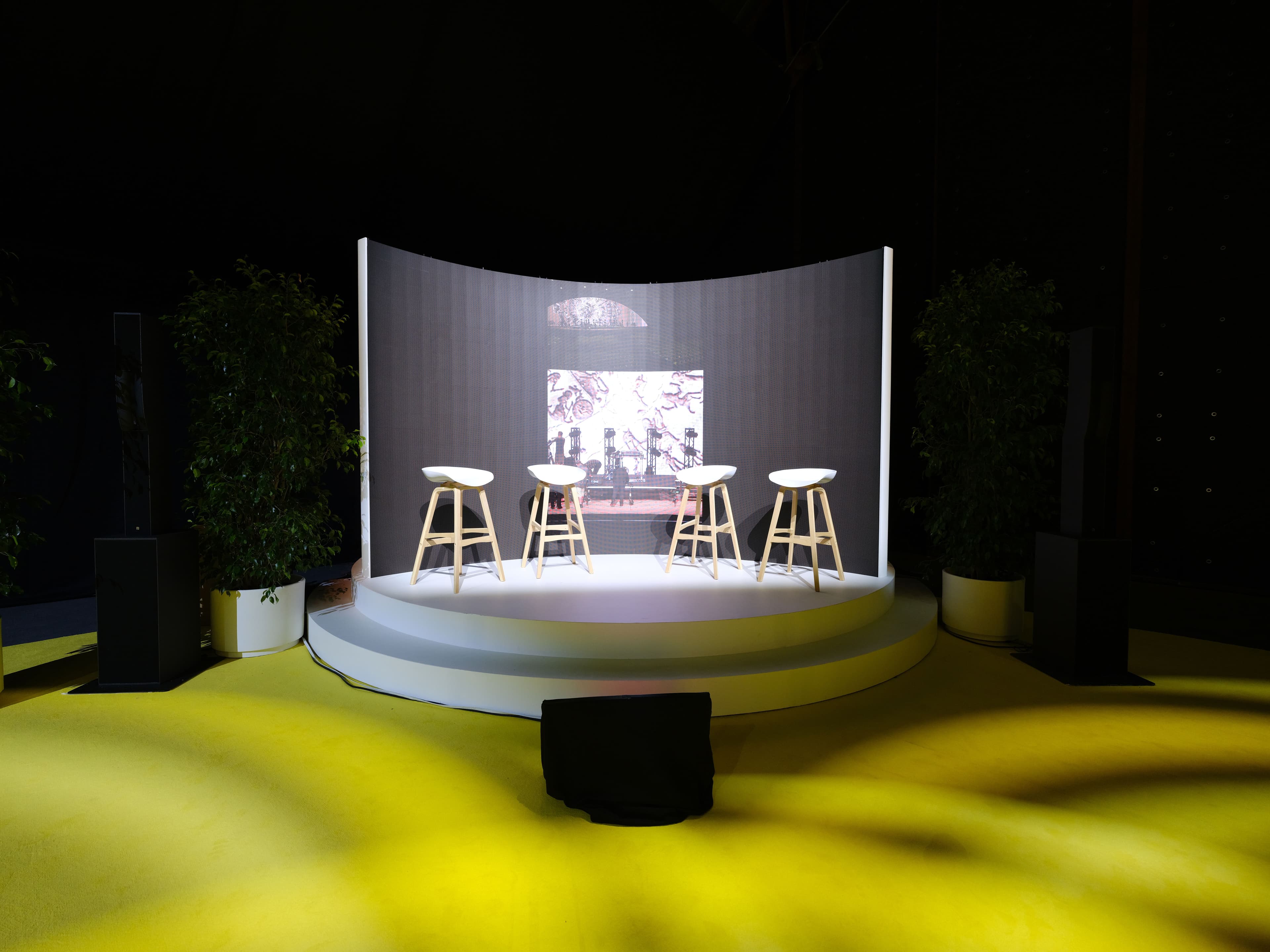 Round yellow stage with stools facing a video screen, lit from below at night.