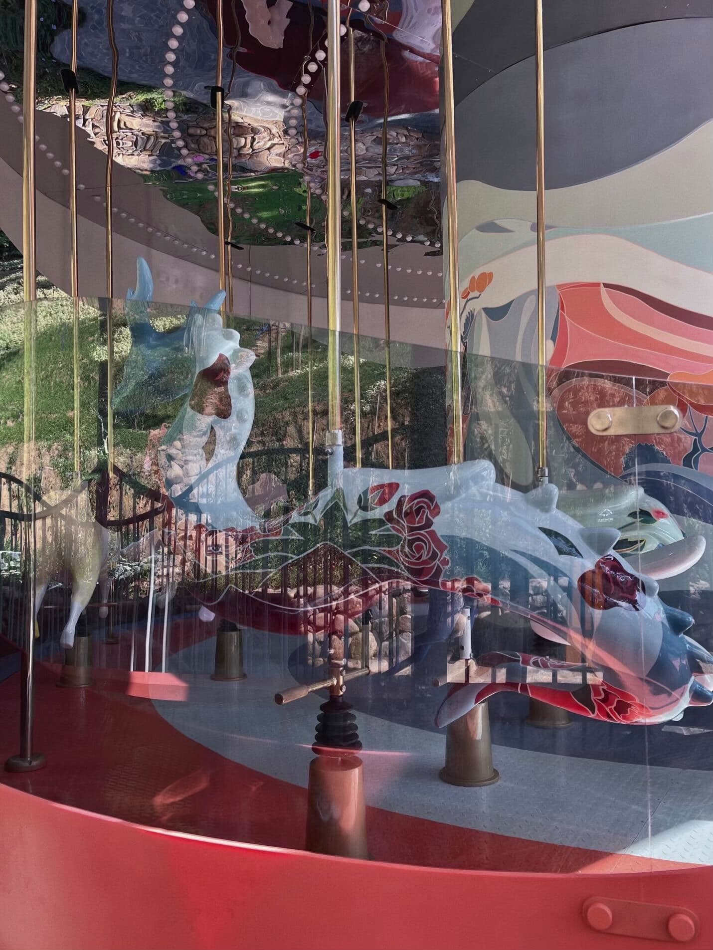 Close-up through carousel canopy glass of illustrated deer and fish figures with gold poles and a painted mural backdrop.