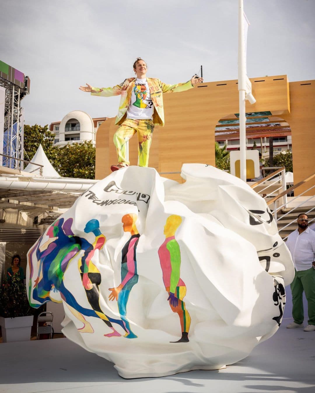 A man in a colorful patterned outfit stands with arms outstretched atop a large crumpled white sculpture painted with colorful figures.