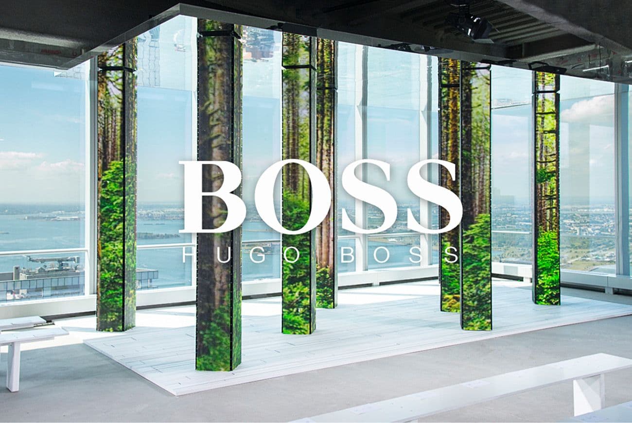 BOSS logo overlaid on a rooftop installation of forest-print LED columns beneath an exposed concrete ceiling, with a harbor and skyline visible through the windows.