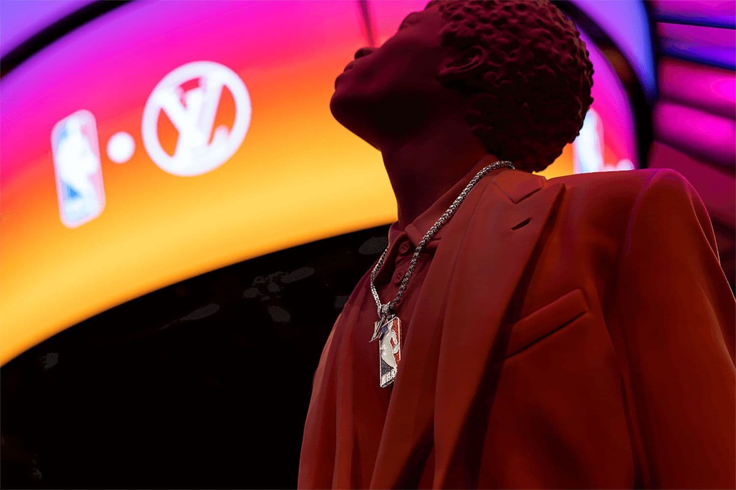 Man in a red suit and layered chain necklace standing before a curved arena video board showing an NBA logo.