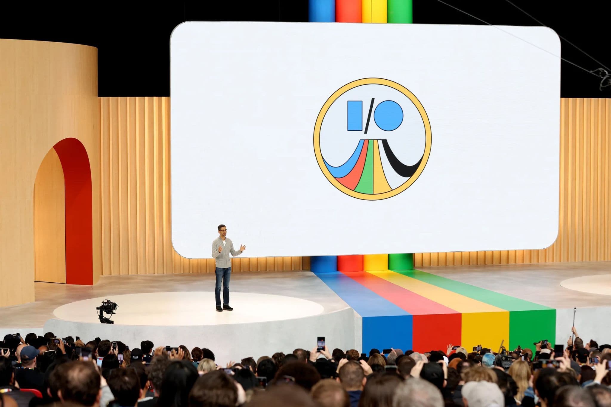 Presenter standing on a circular platform beside a screen showing a circular multicolor I/O emblem, large seated audience.