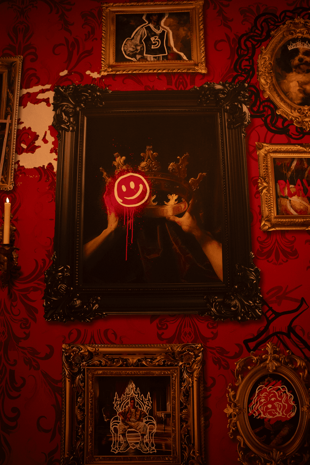 A red gallery wall densely hung with ornate framed artworks, including a painting of hands cradling a glowing red smiley face.