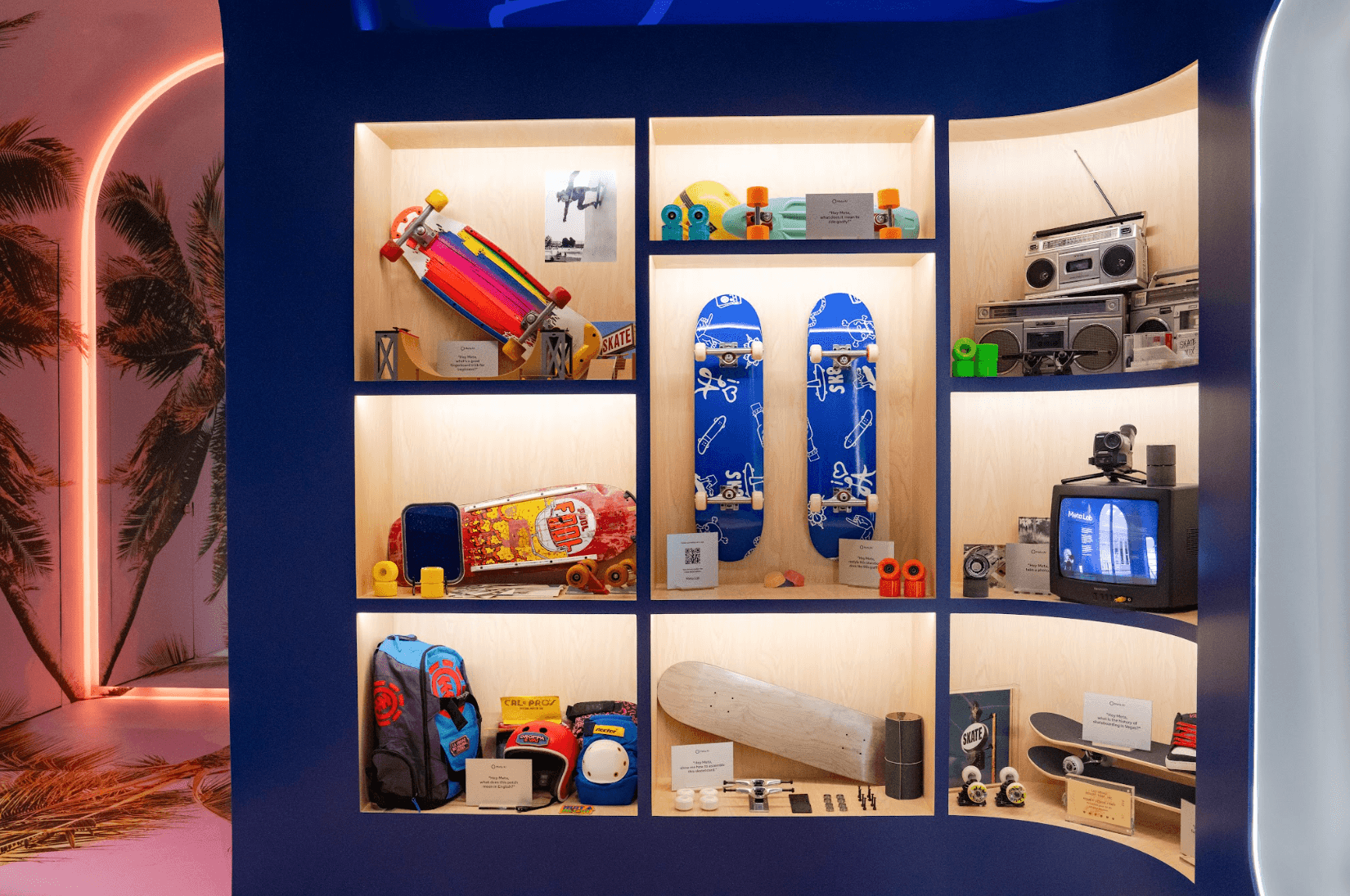 Blue display wall with cubby shelves holding skateboards, a boombox, backpacks, and other lifestyle products, palm motif lighting to the side.