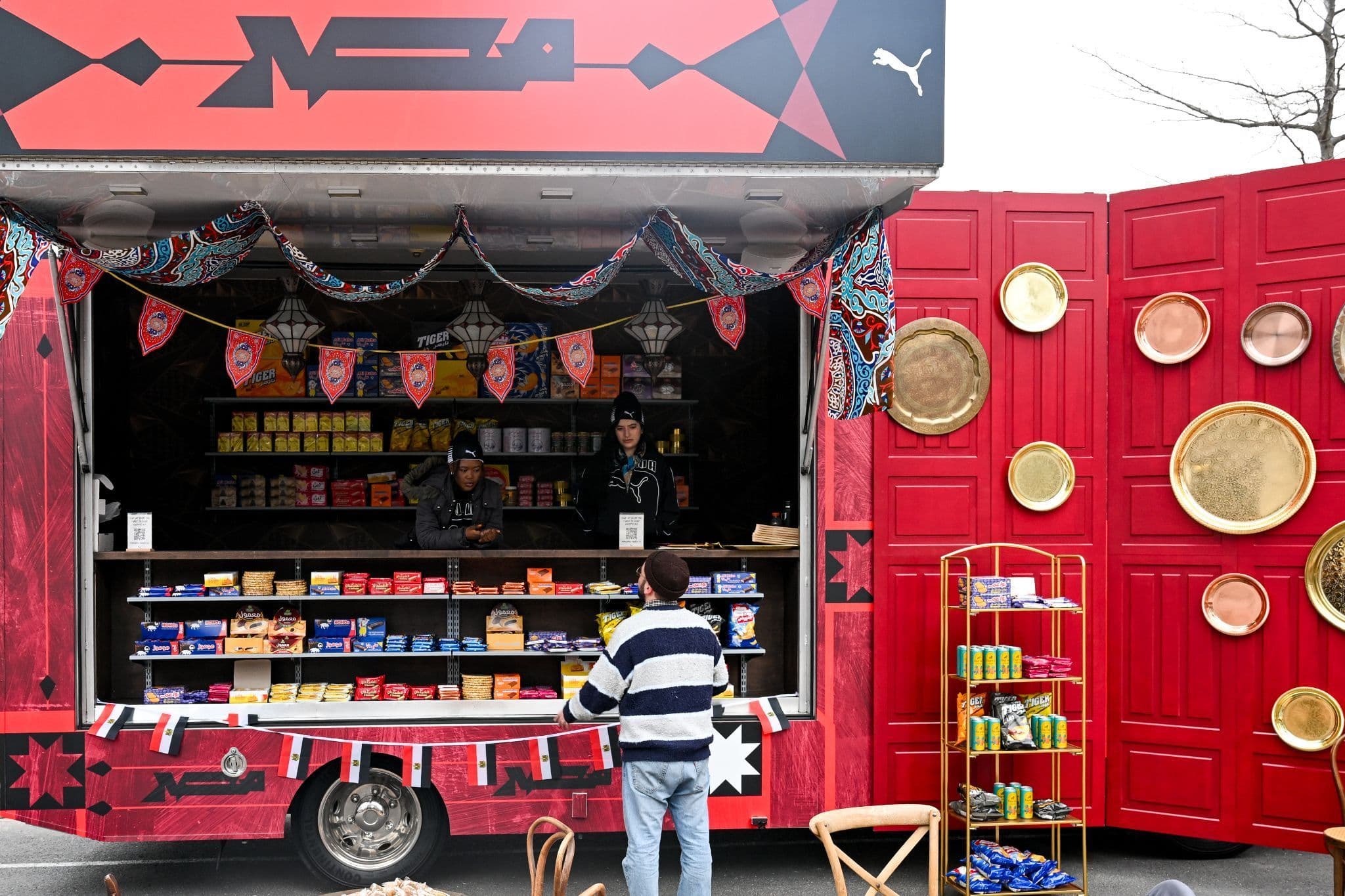 Red shipping-container kiosk with Puma World Cup branding, gold circular displays, and merchandise on shelves, with a shopper browsing inside.