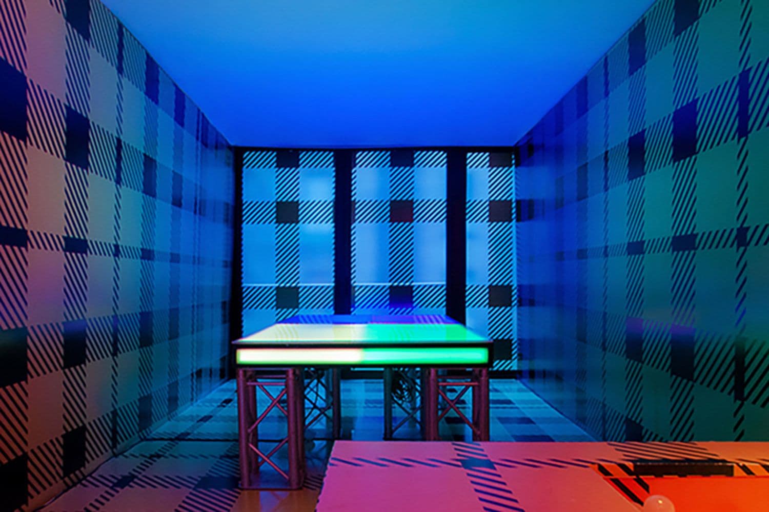 Room with plaid-patterned walls in blue, green and red housing an illuminated ping-pong table.