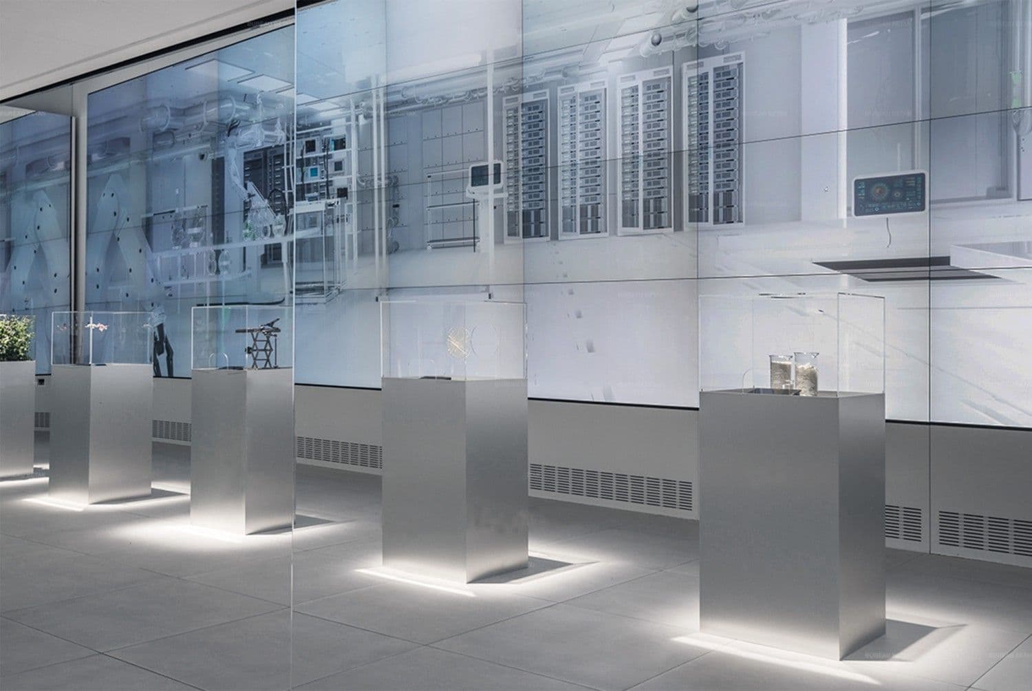 Row of steel pedestals with glass sample cases in front of a large illuminated video wall.