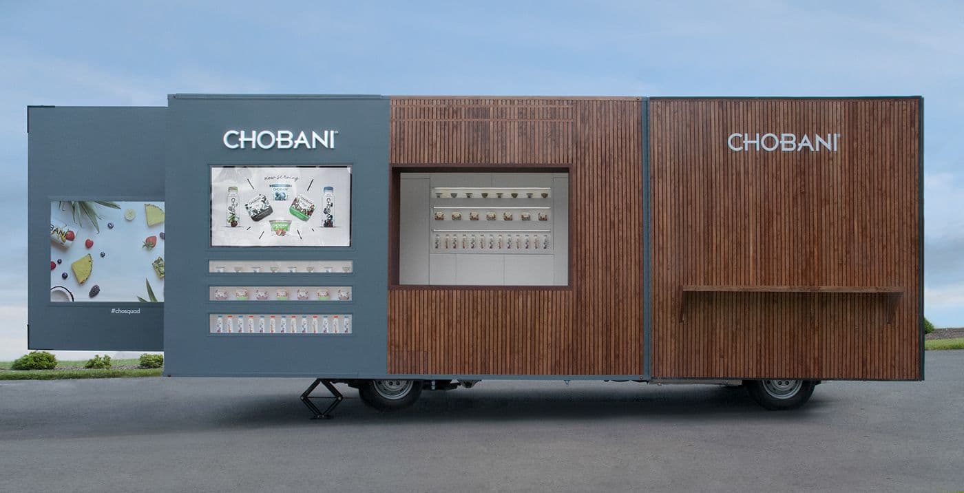 Side view of a wood-slat-clad mobile trailer branded CHOBANI, with a digital menu screen and a vending dispenser built into the facade.