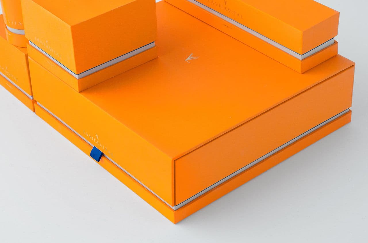 Close-up angled view of stacked orange packaging boxes with a foil-stamped logo and a blue tab pull on the corner.