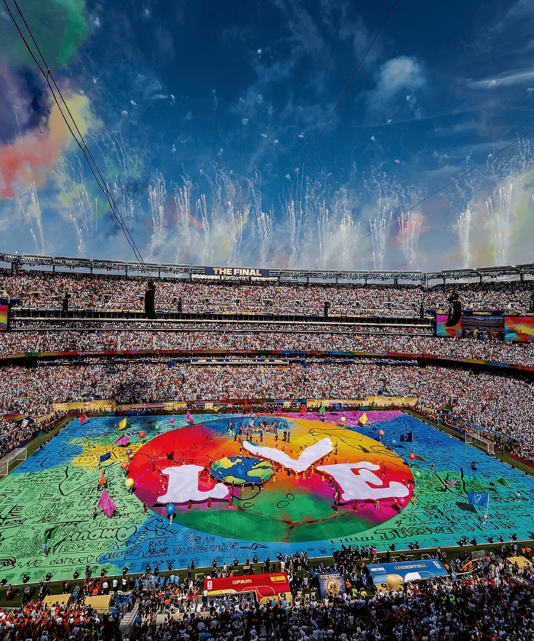 Aerial view of a packed stadium during the World Cup halftime show, with a giant rainbow-colored "LOVE" artwork and dove design covering the field, fireworks and confetti in the sky above the crowd.