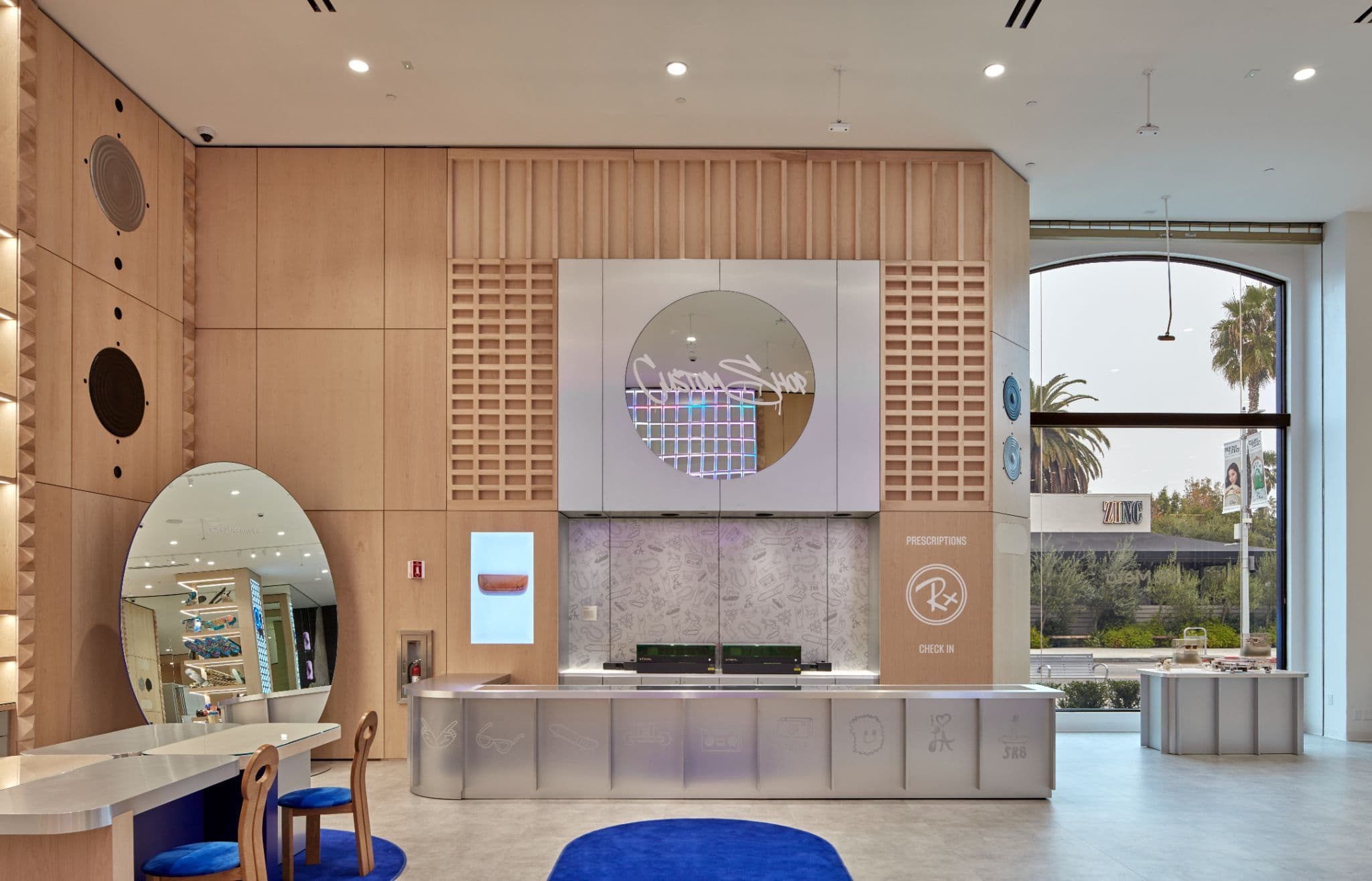 Light wood reception counter with a circular cutout display and round mirror, palm trees visible through the window behind it.