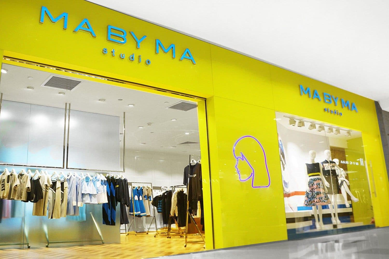 Corner view of the yellow MA BY MA studio storefront with signage, a neon profile logo on the wall, and clothing racks inside.