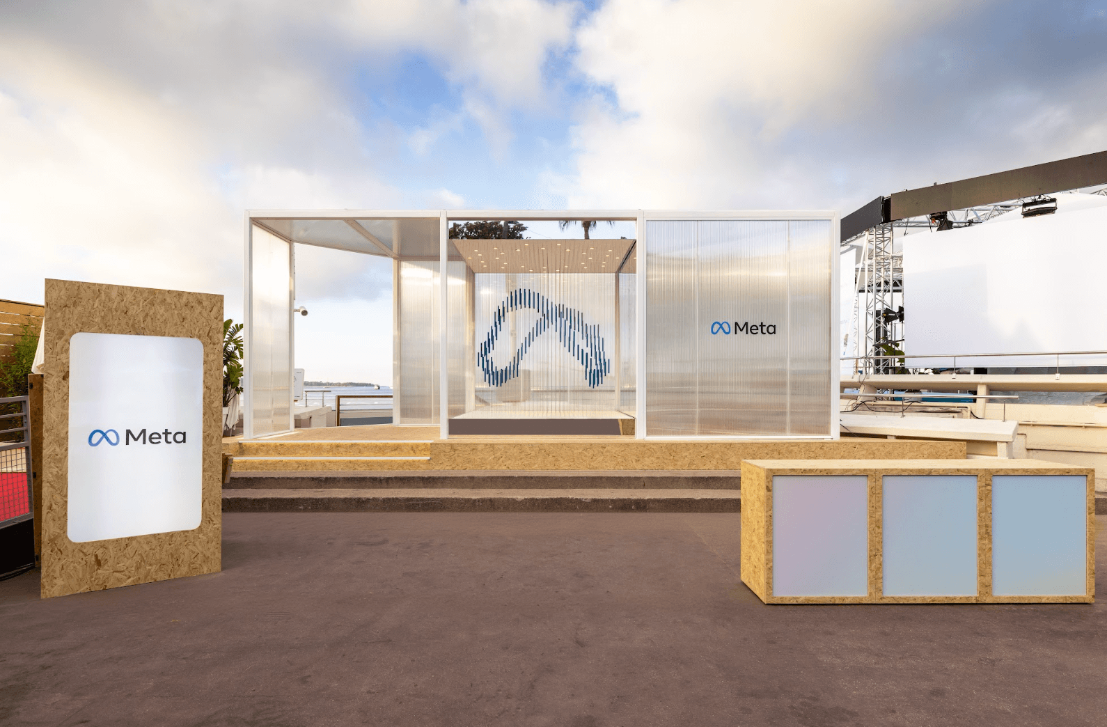 A translucent glass pavilion with a Meta logo on the beachfront, wooden crates and a blue cloud graphic visible inside, under a partly cloudy sky.