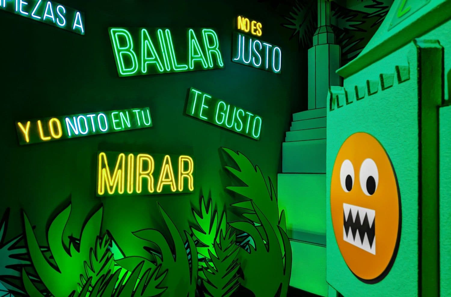 Green neon text reading "Bailar" and "Mirar" beside a glowing orange emoji sculpture.