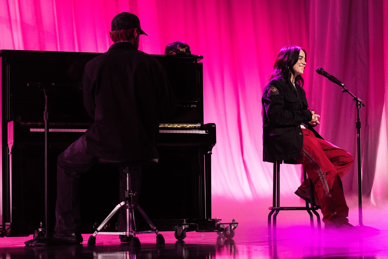 Performer seated at a piano beside a singer on a stool, both lit in pink