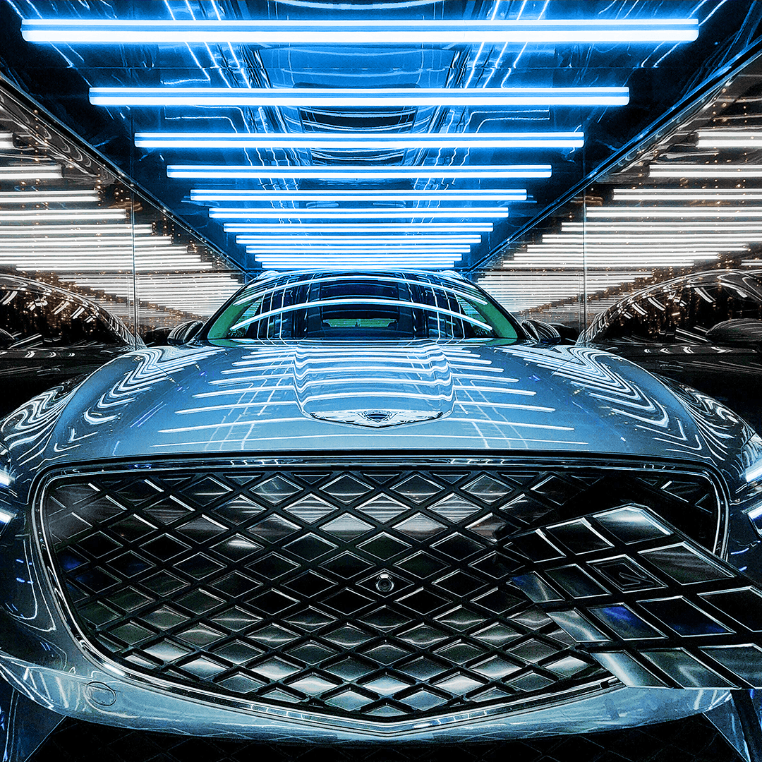 Close-up of a car's front grille and headlights under a mirrored blue neon-tube ceiling, symmetrically reflected.
