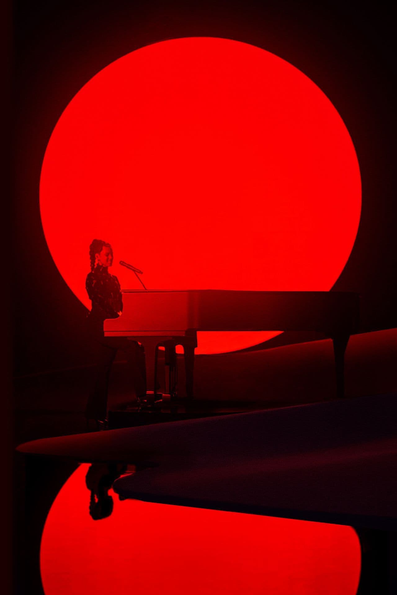 Vertical shot of a performer silhouetted at a piano against a giant red sun and deep red sky, with a red curved dune stage below.