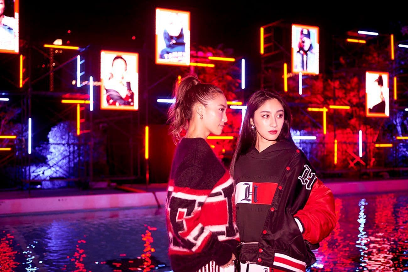 Two women standing beside a reflecting pool at night, backed by a wall of illuminated portrait screens.