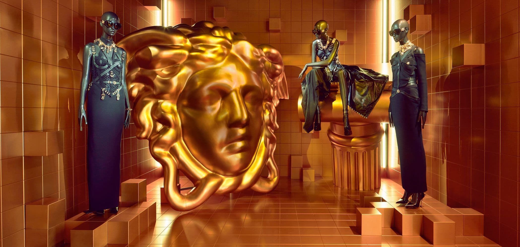 Two mannequins in blue outfits flanking a large gold Medusa head sculpture on a staircase.