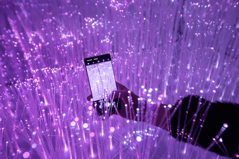A hand holds a smartphone displaying an app screen amid strands of purple fiber-optic light.