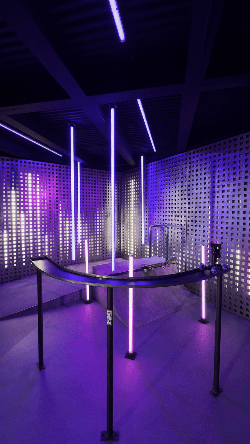 A dark room with purple neon light tubes hanging from the ceiling above a curved black display counter, walls perforated with small square cutouts.